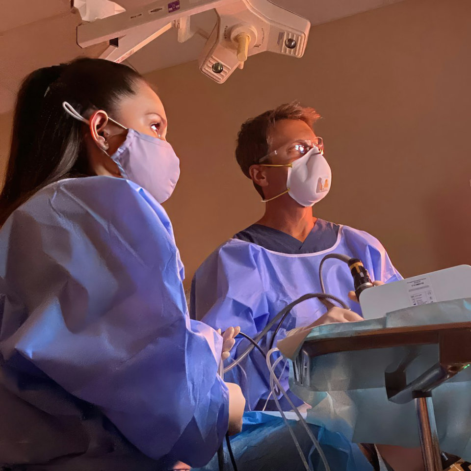 Two healthcare professionals wearing masks and blue gowns operate medical equipment in a clinical setting under overhead lighting.