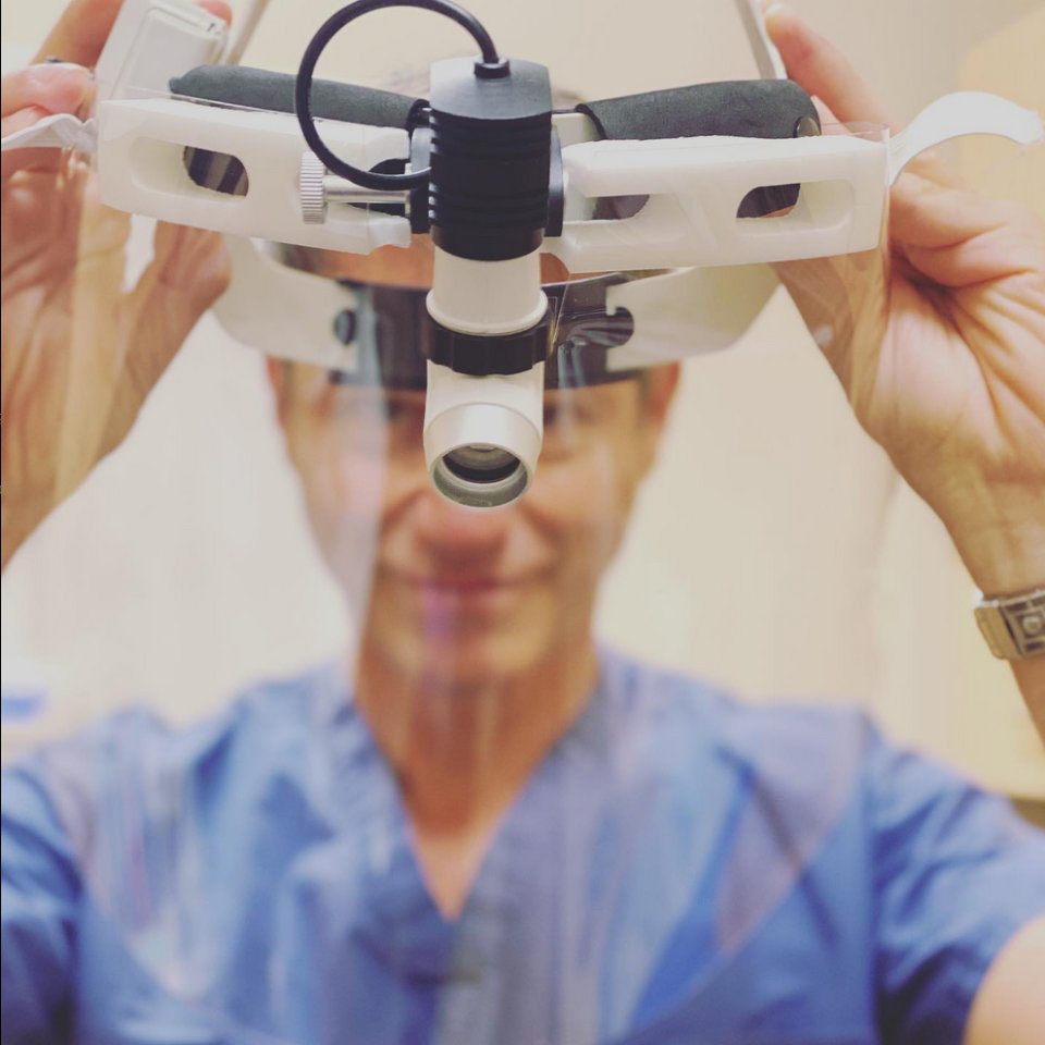 Person in blue scrubs holds up a magnifying medical headlamp with attached face shield, preparing to wear it.