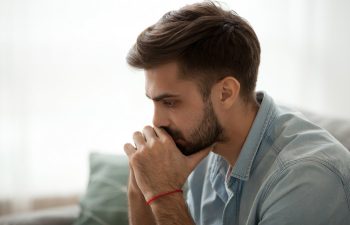 A man with a beard sits indoors, leaning forward with his hands clasped near his mouth, appearing deep in thought.
