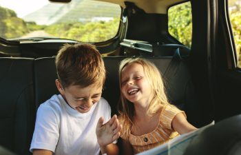 Two young children, a boy and a girl, are laughing together while sitting in the backseat of a car during a daytime road trip.