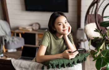 A woman in a green shirt sits indoors, leaning on a cushion, holding a smartphone and smiling at the camera. A laptop and home decor are visible in the background.