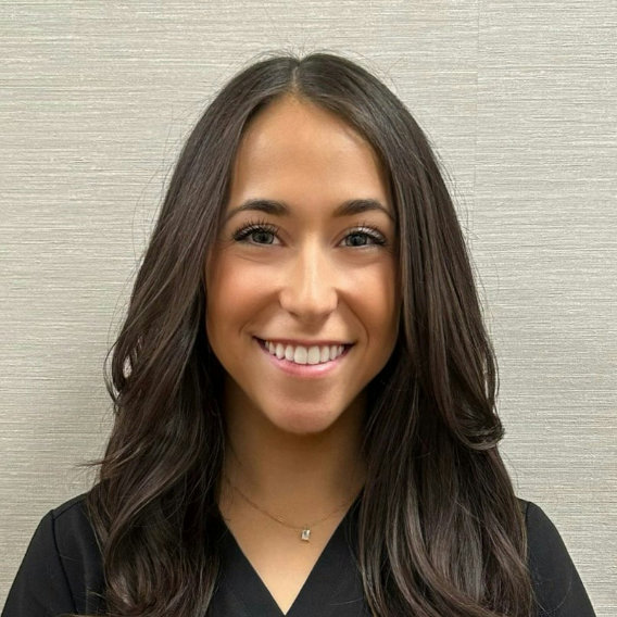 Woman with long, dark hair smiling at the camera, wearing a black top and a necklace, standing in front of a light-colored textured background.