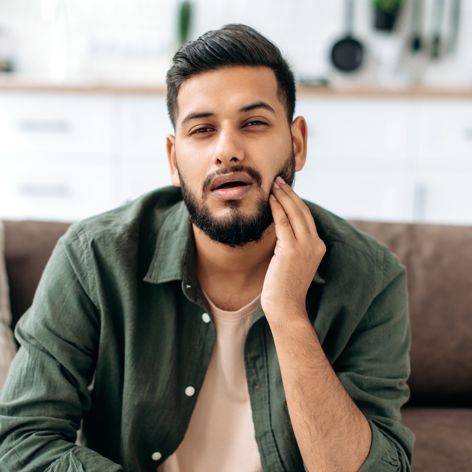 Frustrated unhealthy indian or arabian man in casual clothes, pressing cheek with hand, suffering toothache, caries, sits on a sofa in living room, needs the help of a dentist. Dental problems concept