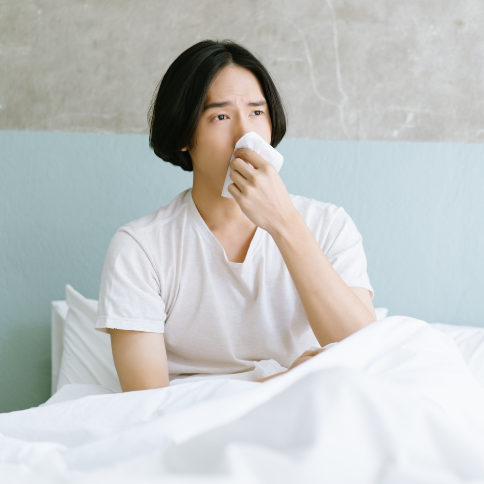 Young Asian man sitting on bed holding tissue handkerchief blowing running nose feels unwell unhealthy. Sick man having influenza symptoms coughing at home. Seasonal allergy or cold fever flu concept.