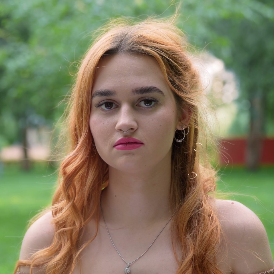 A beautiful red-haired girl with a serious face looks at the camera in nature in summer