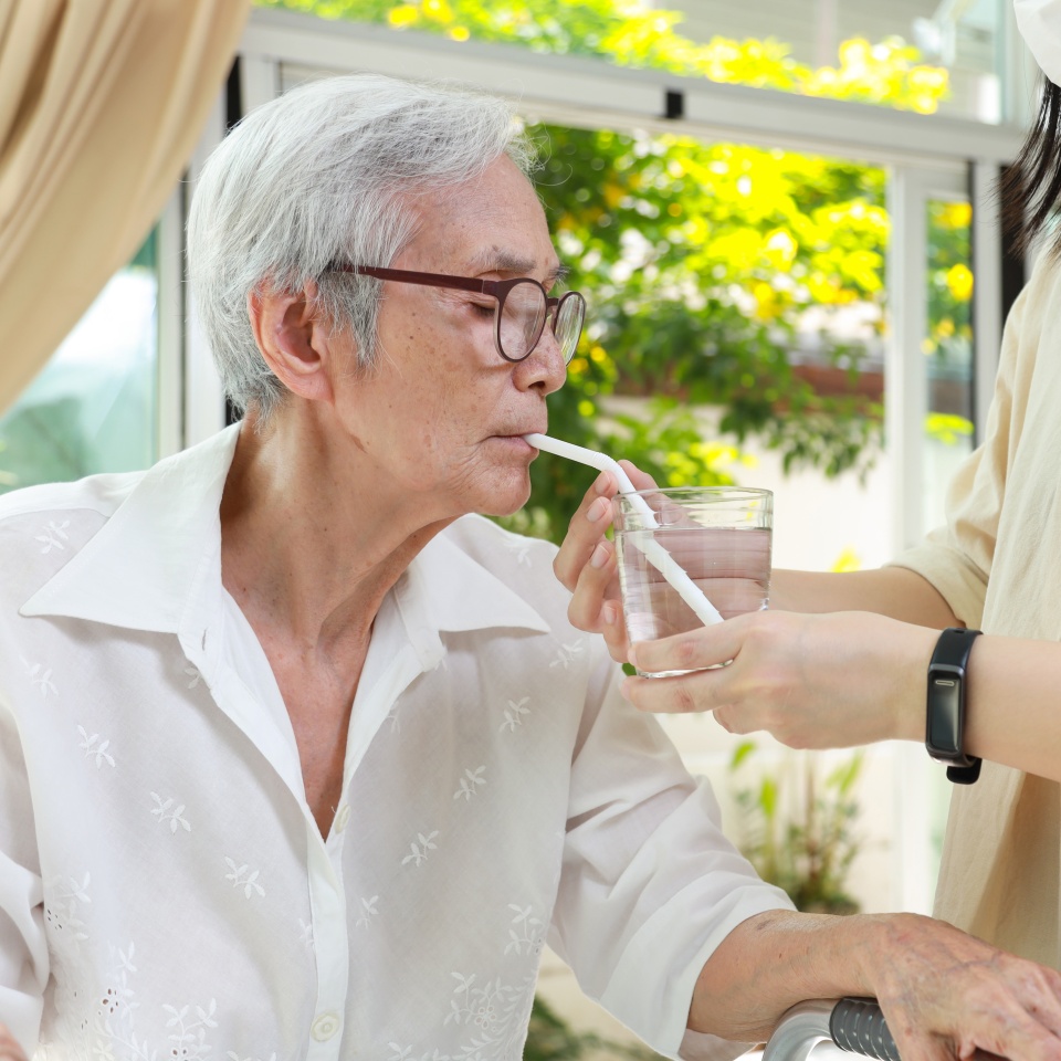 Asian caregiver take care of senior woman give drinking water from a glass,feeding water using a straw to prevent choking at nursing home,thirsty elderly patient sucking water through straw for safety