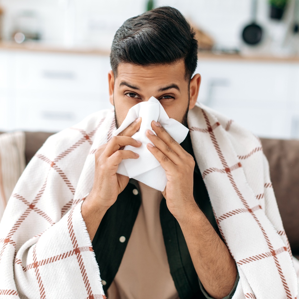 Sneezing, runny nose, cough. Unhealthy sad arabian or indian young man, sits on the couch at home under a plaid, sneezing in a napkin, feels weak, unwell, needs treatment and medicines