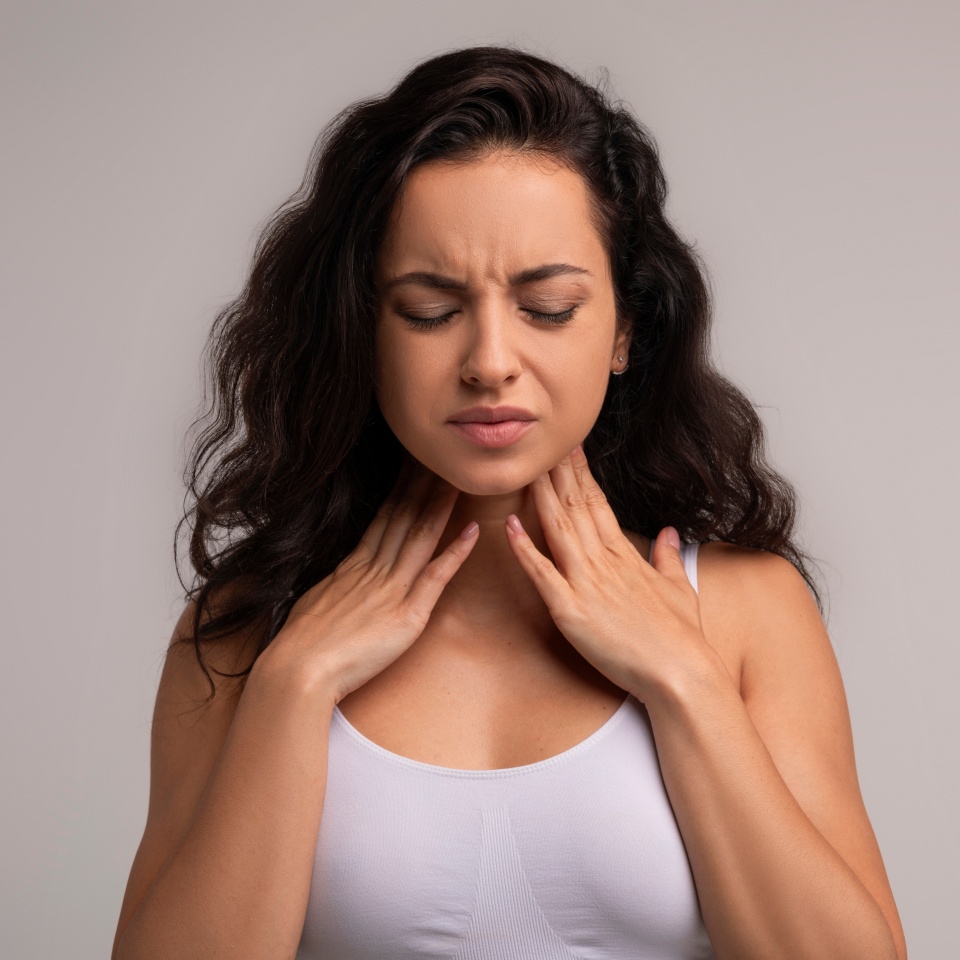Sore throat, tonsilitis concept. Unhappy millennial brunette woman touching her neck, suffering from pain in throat, checking enlarged adenoids isolated on grey studio background, copy space