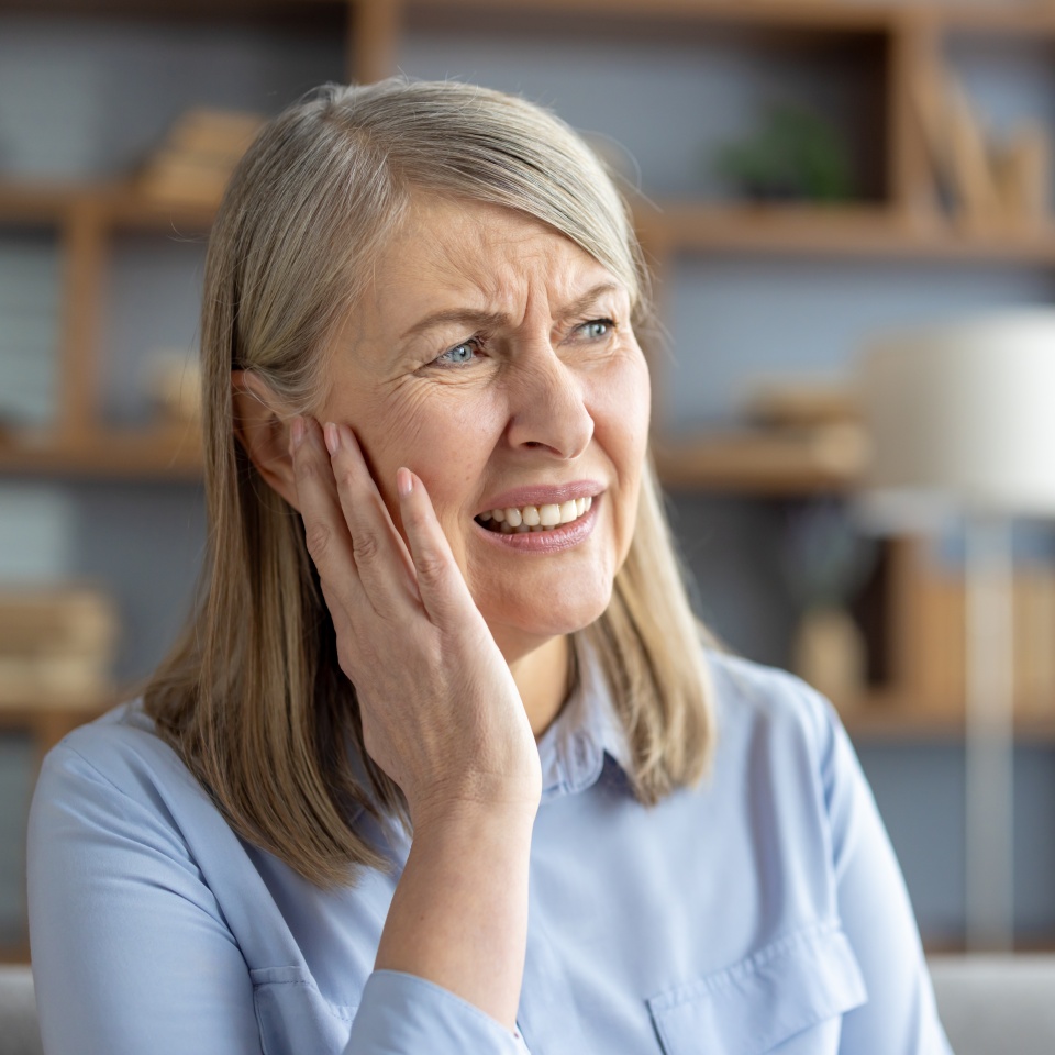 Elderly Caucasian woman suffering from otitis, touching her ear in pain, sitting in a modern living room. She looks distressed and worried about her earache.
