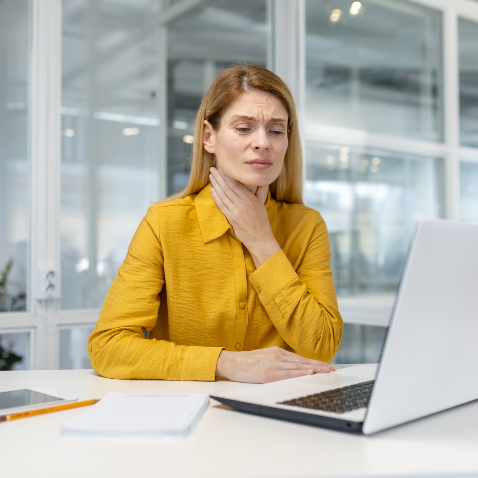 A professional woman in a bright yellow blouse looks uncomfortable and holds her throat while working on a laptop in a bright, airy office.