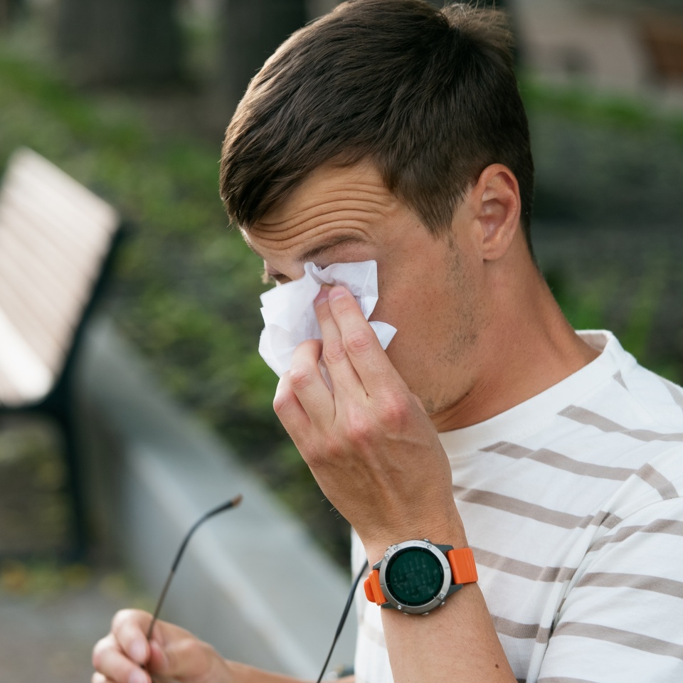 Young man wiping watery eyes with a tissue while holding glasses in a park, experiencing discomfort from seasonal allergies and the effects of pollen in the air