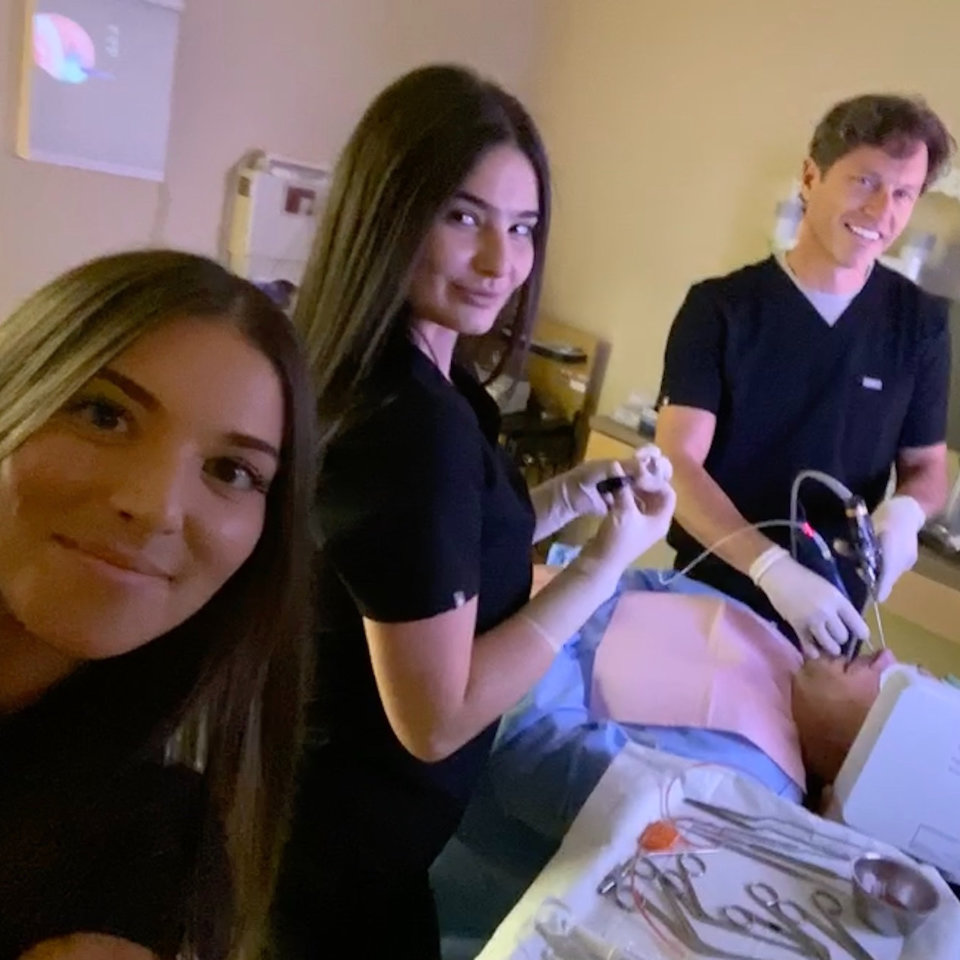 Three medical professionals in scrubs perform a procedure on a patient lying on a hospital bed, with surgical instruments visible in the foreground.