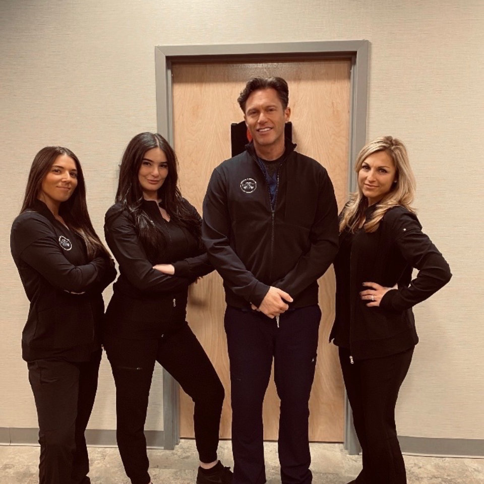 Four people wearing black uniforms stand in front of a closed wooden door, posing and smiling for the camera indoors.