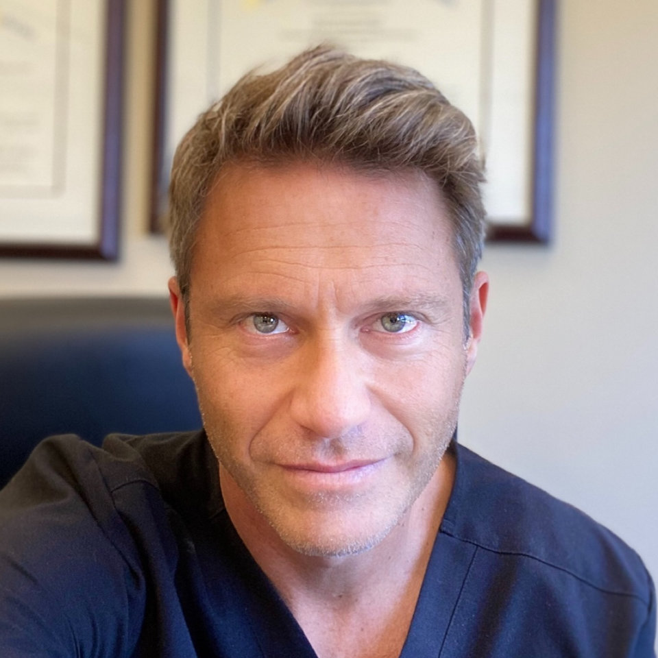 A man with short, light brown hair and light eyes is sitting indoors, wearing dark scrubs. Framed documents hang on the wall behind him.