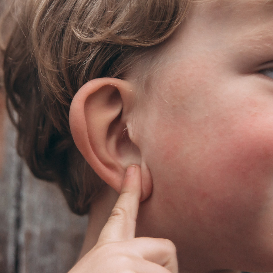 Close-up of a child with light brown hair touching their ear with their index finger.