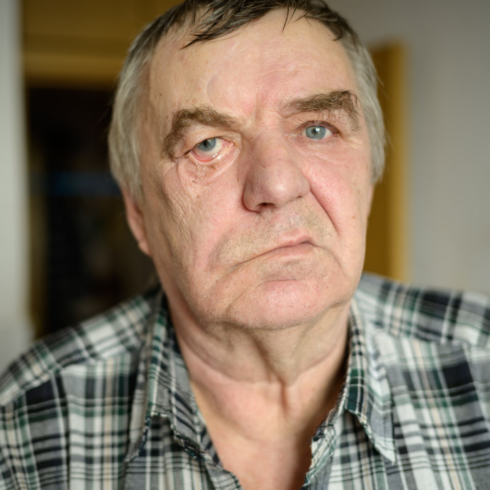 Older man with gray hair and blue eyes wearing a plaid shirt, looking directly at the camera with a neutral expression.