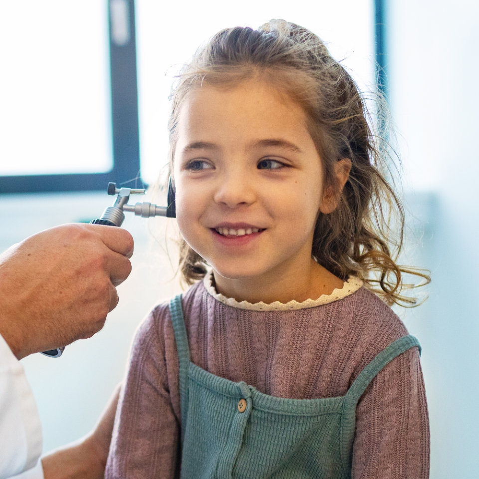 A child sits and smiles while a healthcare professional examines her ear with an otoscope in a brightly lit room.