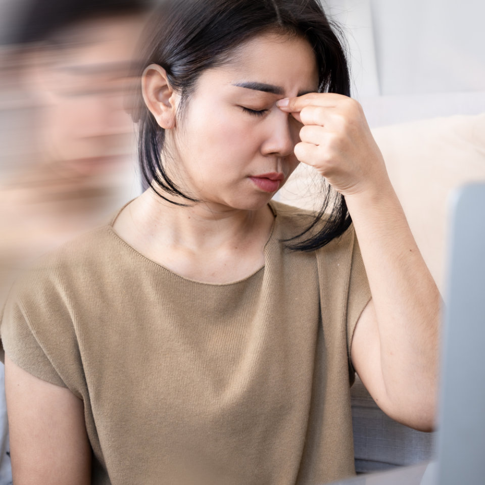 A woman sits with her eyes closed, pinching the bridge of her nose, appearing stressed or fatigued. A blurred duplicate image suggests dizziness or mental strain.
