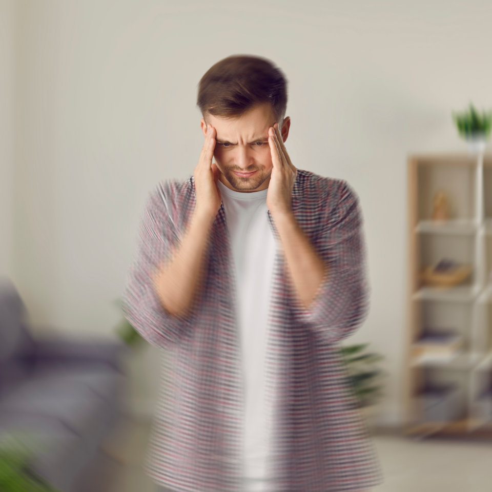 A man stands indoors with his hands on his temples and eyes closed, appearing stressed or in pain. The background is blurred, suggesting dizziness or discomfort.