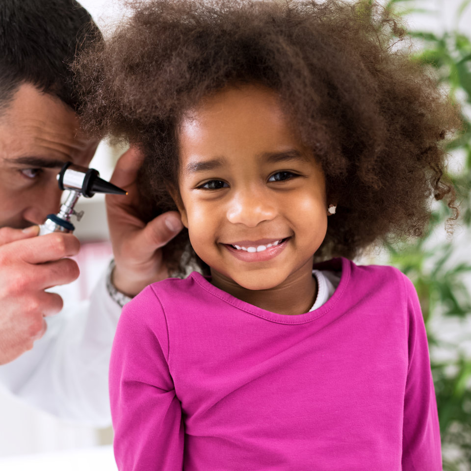 A doctor examines a smiling young girl's ear with an otoscope. The girl is wearing a pink shirt and looking at the camera.