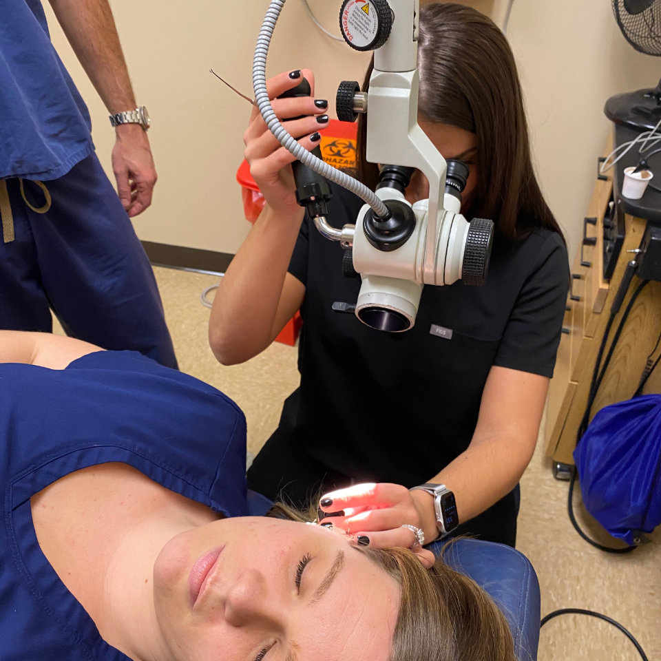 A healthcare professional examines a patient’s ear with a microscope and light while the patient lies on an exam chair.