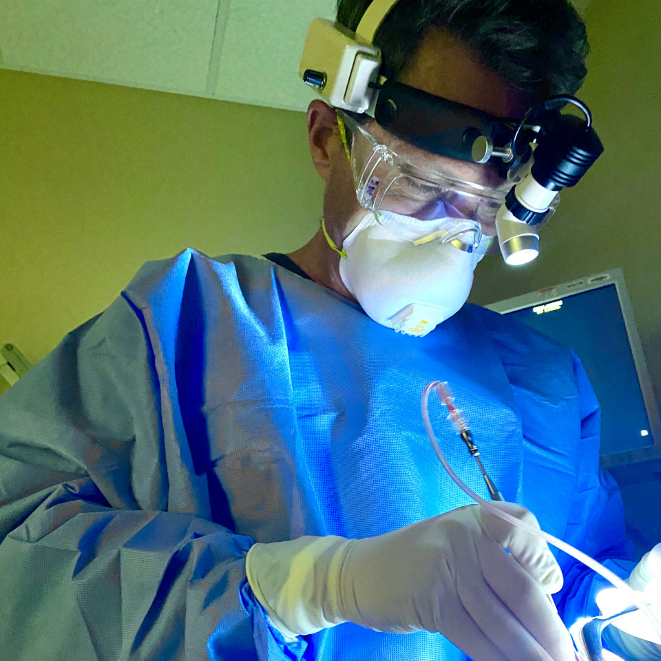 A medical professional in protective gear and face mask performs a procedure under bright light, using surgical instruments.
