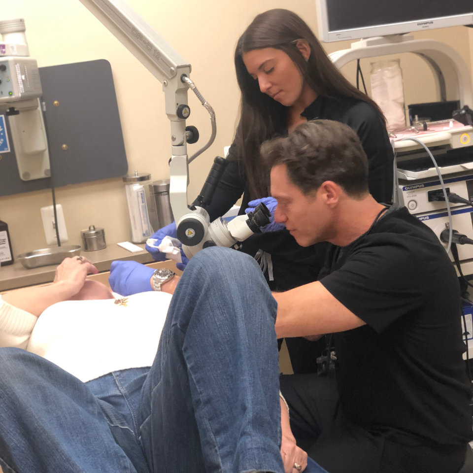 A man uses a microscope to examine a patient lying on an exam table, while a woman assists in a medical office setting.