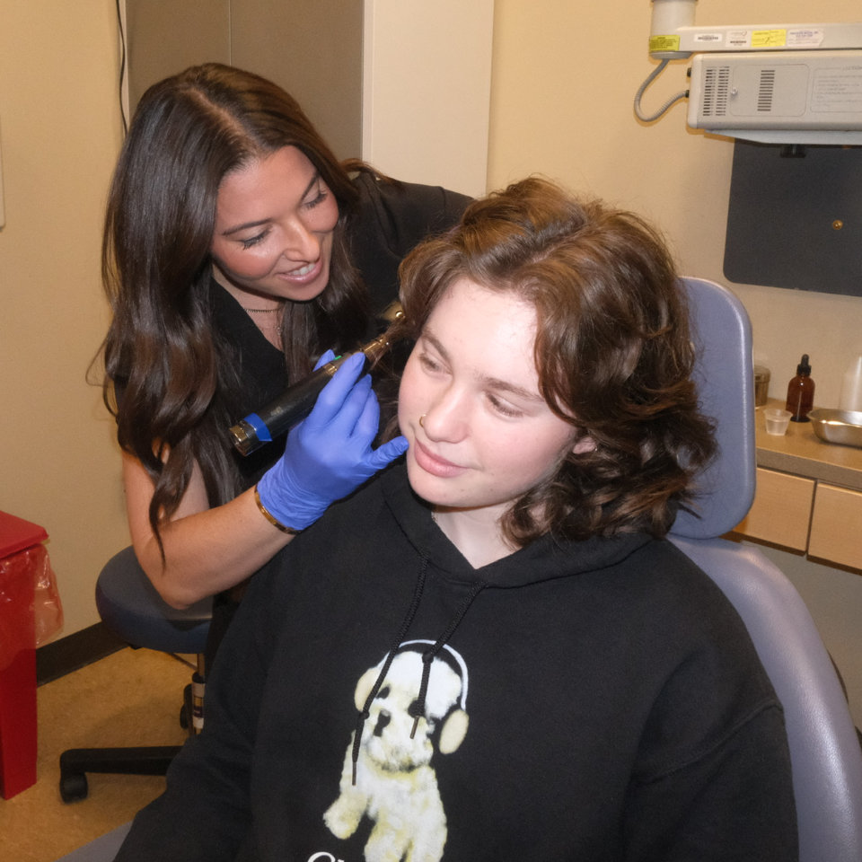 A healthcare professional uses a handheld device on a patient's cheek while the patient sits in a clinic chair.