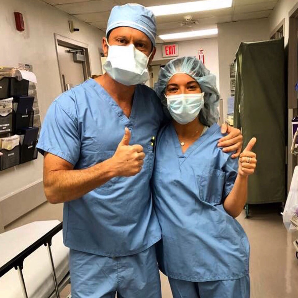 Two people in blue surgical scrubs, masks, and caps stand in a hospital hallway, giving thumbs up and posing for the camera.