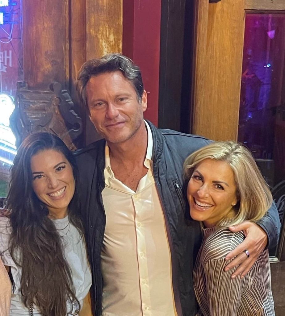 Three adults, two women and one man, stand closely together indoors, smiling at the camera. Wood paneling and dim lighting are visible in the background.