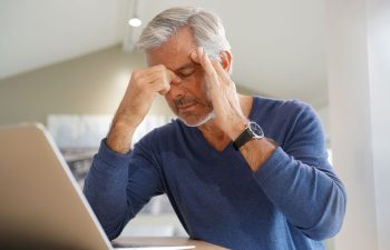 An older man sits at a desk with a laptop, eyes closed and fingers on his temples, appearing to have a headache caused by sinusities and feel fatigued.